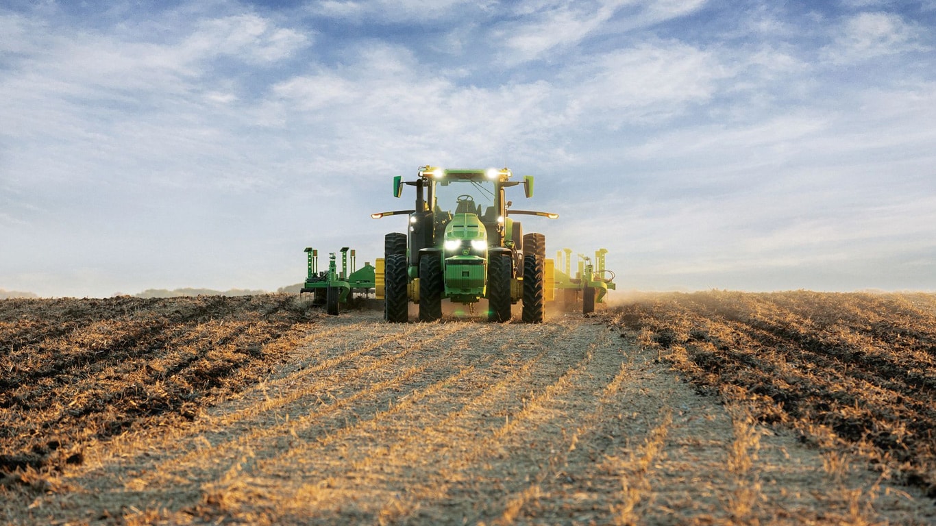 Selbstfahrender John Deere Traktor zieht Bodenbearbeitungsgeräte durch ein offenes Feld.
