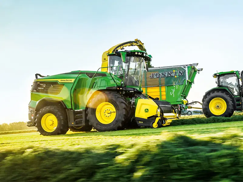 John Deere F8&nbsp;600 Feldhäcksler sammelt Gras neben einem Traktor und Feldanhänger auf dem Feld